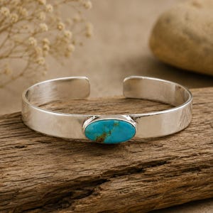 May include: A silver cuff bracelet featuring a smooth, polished band with an open back design. Centered on the front is a single, oval-shaped turquoise stone displaying a vibrant blue color with natural brown matrix veining throughout.