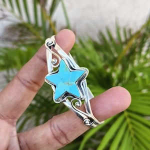 May include: A silver cuff bracelet featuring a central star-shaped turquoise stone with natural brown matrix inclusions. The metal band is crafted with polished silver wirework, showcasing decorative swirling patterns that frame the star centrepiece.