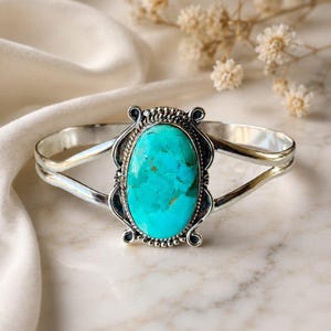May include: A silver cuff bracelet featuring a large, oval-shaped turquoise stone at the centre. The stone is bright blue with natural dark veining. The metal setting has a decorative beaded border and a split band design on both sides.