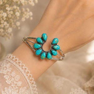 Könnte beinhalten: Ein silbernes Armband mit einem hufeisenförmigen Mittelstück, das mit neun tropfenförmigen Türkissteinen besetzt ist. Das Metallband hat eine texturierte, oxidierte Oberfläche und die Steine zeigen leuchtend blaue Farbtöne mit braunen Maserungen.