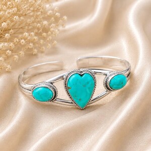 May include: A silver cuff bracelet featuring a central heart-shaped turquoise stone flanked by two smaller oval turquoise stones. The metal band has a twisted rope texture bordering each stone setting, creating a detailed and polished appearance.