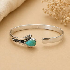 May include: A silver cuff bracelet featuring a teardrop-shaped turquoise stone set at one end. The metal band has a polished finish with oxidised detailing along the side, including a textured rope-like pattern near the stone setting.