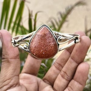 Puede incluir: Un brazalete de plata con una gran piedra de sol en forma de pera en el centro. La banda de metal presenta detalles intrincados de alambre con pequeñas esferas de plata a cada lado de la piedra, creando un marco decorativo abierto.