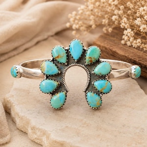 May include: A silver cuff bracelet featuring a central horseshoe-shaped element adorned with nine teardrop-shaped turquoise stones. The stones display vibrant blue hues with brown matrix veining. Two additional turquoise stones are set on the band.