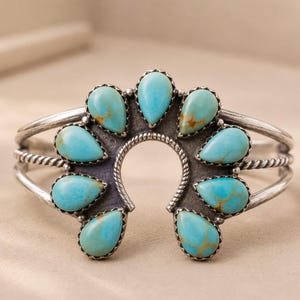 May include: A silver cuff bracelet featuring a central horseshoe-shaped element adorned with eight teardrop-shaped turquoise stones. The metal has a darkened, oxidised finish, and the band consists of two parallel wires with a twisted rope texture.