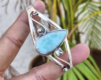 May include: A silver cuff bracelet featuring a central teardrop-shaped light blue Larimar gemstone. The metal band is crafted with openwork swirling wire designs that frame the stone, creating a decorative and polished metallic finish.
