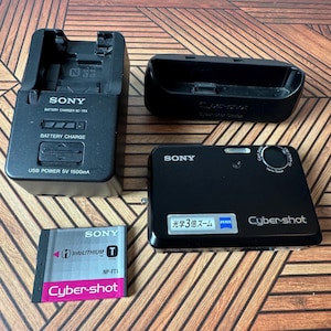 May include: A black Sony Cyber-shot digital camera with a Zeiss lens, a Sony BC-TRX battery charger, a black docking station, and a Sony InfoLithium T-type NP-FT1 battery. The camera features a label with Japanese text and a Zeiss logo.