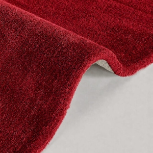 Puede incluir: Una alfombra de color rojo intenso con un pelo grueso, afelpado y de textura suave. La alfombra está doblada para revelar un vistazo del material de soporte tejido de color claro debajo de las densas fibras de color carmesí.