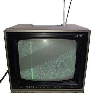 Könnte beinhalten: Ein alter Hitachi-Fernseher mit grauem Kunststoffgehäuse und einem schwarzen Bildschirm, der Rauschen anzeigt. Das Gerät verfügt über zwei ausziehbare Metallantennen, ein frontseitiges Lautsprechergitter, manuelle Drehregler und Schalter.