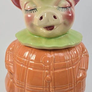 May include: A ceramic cookie jar shaped like a pig wearing a green hat and a light green ruffled collar. The pig has closed eyes with long eyelashes, pink cheeks, and a smiling mouth. The body is orange with a raised plaid pattern and buttons.