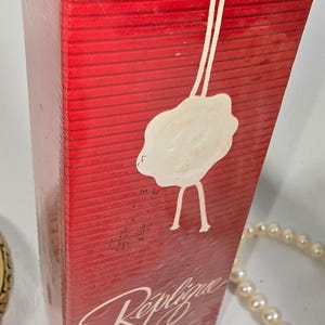 May include: A rectangular red perfume box with horizontal lines, featuring a white wax seal emblem on the front. The text reads Replique Eau de Toilette SPRAY 2 FL. OZ 60 ML. The box is wrapped in clear plastic and has a white string detail.
