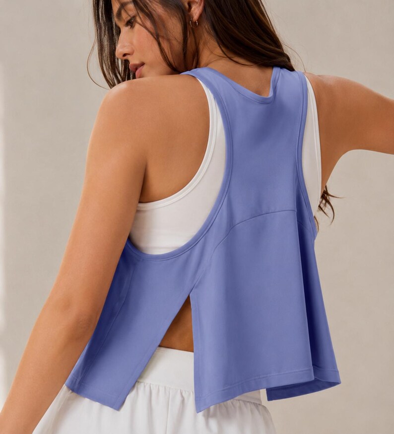 May include: A periwinkle blue athletic tank top with a racerback design and deep side slits, layered over a white sleeveless base layer. The top features a cropped, loose-fitting silhouette with wide armholes and a smooth, solid-colored fabric finish.