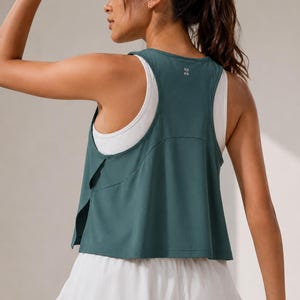 May include: A dark teal racerback tank top layered over a white tank top, paired with a white tiered skirt. The teal top features a split side design and a small reflective logo on the upper back. The outfit is designed for athletic movement.