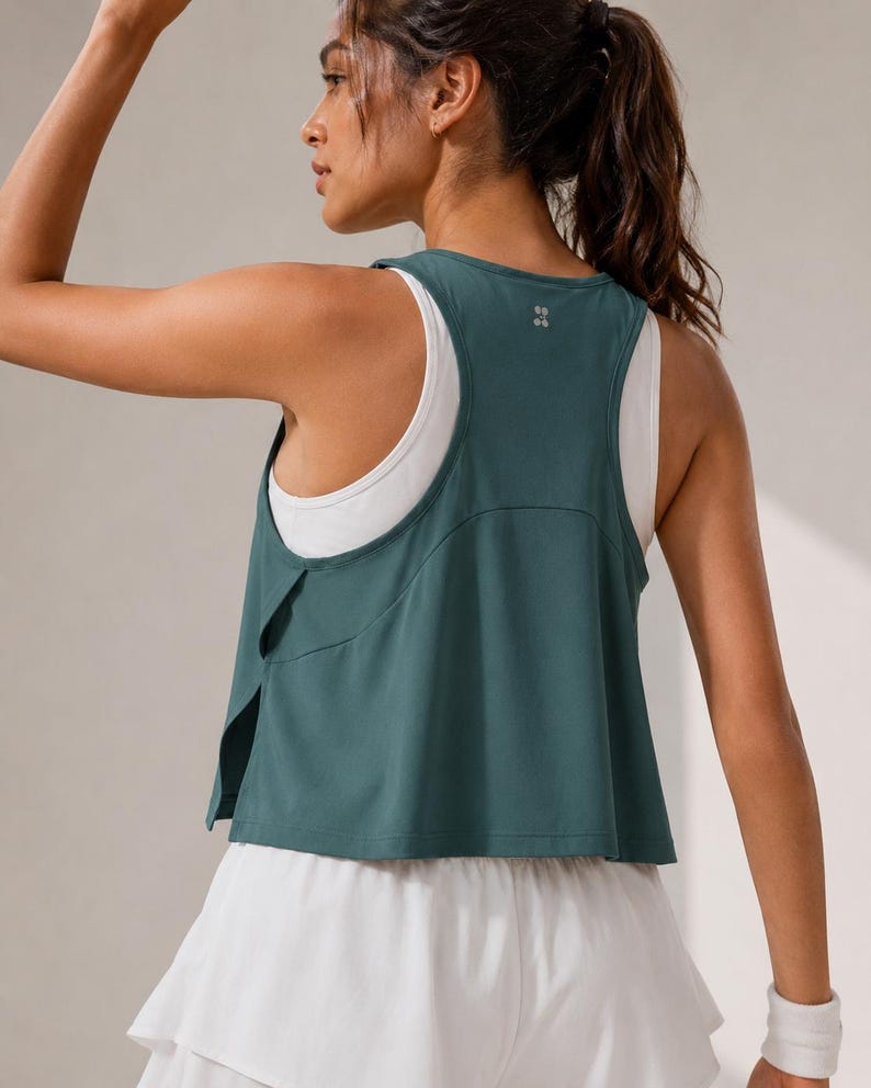 May include: A dark teal racerback tank top layered over a white tank top, paired with a white tiered skirt. The teal top features a split side design and a small reflective logo on the upper back. The outfit is designed for athletic movement.