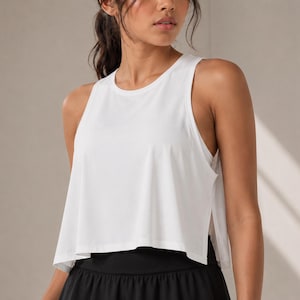 May include: A solid white sleeveless tank top featuring a crew neckline, a loose cropped fit, and deep side slits that reveal a layered hemline. The garment is constructed from a lightweight, smooth fabric with a clean, minimalist aesthetic.