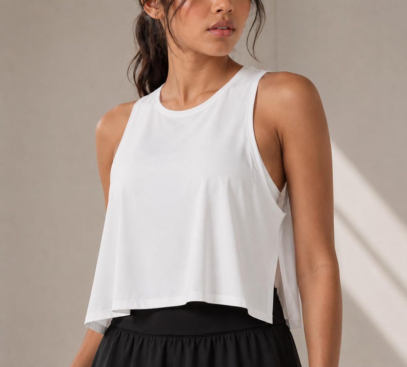 May include: A solid white sleeveless tank top featuring a crew neckline, a loose cropped fit, and deep side slits that reveal a layered hemline. The garment is constructed from a lightweight, smooth fabric with a clean, minimalist aesthetic.