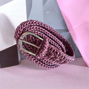 Op de afbeelding: Een gehaakte riem in de kleur mauve met een gestructureerd, gevlochten patroon over de gehele band. De riem wordt vastgezet met een gepolijste, zilverkleurige metalen gesp met een afgerond, halfrond frame en een enkele doornsluiting.