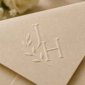 May include: A cream-colored envelope featuring a blind embossed design on the flap. The design consists of the capital letters J and H in a serif font, accompanied by a delicate, embossed sprig of leaves curving along the left side of the letters.