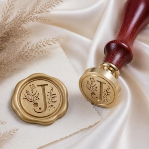 May include: A wax seal stamp with a dark reddish-brown wooden handle and a circular brass head featuring the letter J surrounded by floral engravings, accompanied by a matching gold-coloured wax seal impression on a piece of cream-coloured paper.