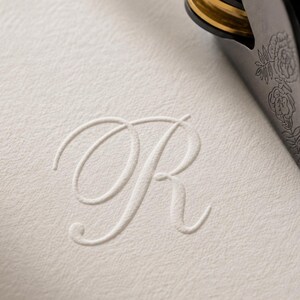 May include: A white textured paper surface featuring a debossed cursive letter R in the center. A portion of a dark metal embossing tool with a gold-colored ring and an engraved floral pattern is visible in the upper right corner of the frame.
