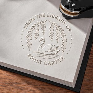 Może przedstawiać: Okrągły odcisk tłoczonego stempla na białym papierze przedstawiający łabędzia pod gałęziami wierzby. Tekst FROM THE LIBRARY OF EMILY CARTER znajduje się w okrągłej ramce. Czarne metalowe urządzenie do tłoczenia jest widoczne w prawym górnym rogu.