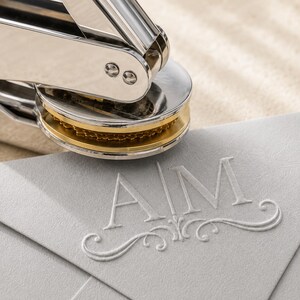 May include: A silver metal hand-held embosser tool with a circular gold-toned die plate pressing onto a light gray paper envelope. The embossed impression on the paper features the capital letters AIM centered above a decorative flourish design.