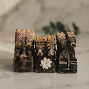 May include: Three pairs of carved wooden horse figurines arranged in a row. Each pair features dark, weathered wood with hand-painted details in red, white, and yellow. A small, white, multi-petaled flower is placed on the center pair of horses.