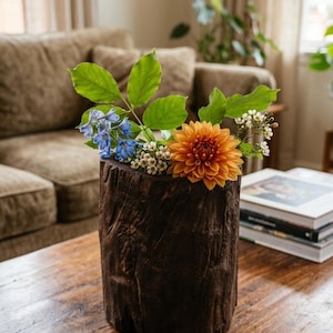 Puede incluir: Un florero cilíndrico hecho de corteza de madera de color marrón oscuro con textura. El florero contiene un arreglo floral que presenta una dalia naranja grande, pequeños racimos de flores blancas, espuelas de caballero azul brillante y varias hojas verdes.