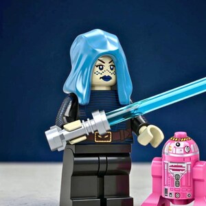 May include: A plastic minifigure with a light blue hood, dark blue lipstick, and a blue lightsaber, wearing a dark blue and black outfit with a brown belt. Beside the minifigure stands a small pink and white droid with printed mechanical details.