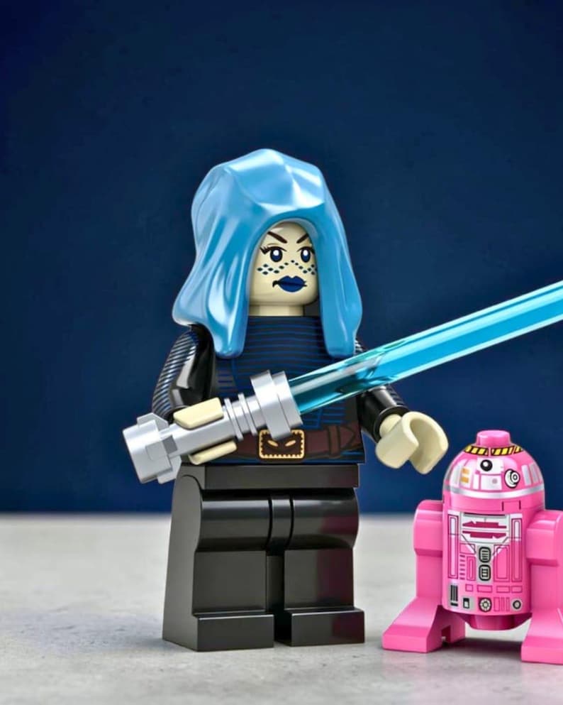May include: A plastic minifigure with a light blue hood, dark blue lipstick, and a blue lightsaber, wearing a dark blue and black outfit with a brown belt. Beside the minifigure stands a small pink and white droid with printed mechanical details.