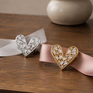 Op de afbeelding: Twee hartvormige decoratieve accenten met heldere baguette- en ronde kristallen in een metalen zetting. Het ene hart is zilverkleurig op een lichtgrijs lint, het andere goudkleurig op een oudroze lint, beide met een gefacetteerd ontwerp.