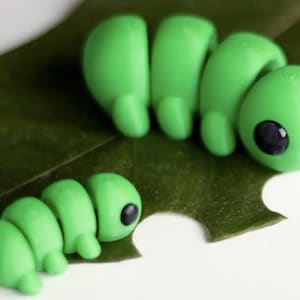 May include: Two green caterpillar figurines with black eyes rest on a green leaf. The caterpillars are segmented, with rounded body sections. One caterpillar is positioned below the other, with a white background.