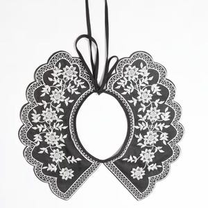 May include: A black detachable collar featuring white floral embroidery and a scalloped edge. The collar has a rounded shape with a central opening and includes two thin black ribbon ties at the top for securing the garment around the neck.