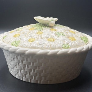 May include: A round ceramic lidded dish with a white basket-weave texture on the base and a braided rim. The lid features a raised relief pattern of white daisies with yellow centers and green leaves, topped with a small sculpted butterfly handle.