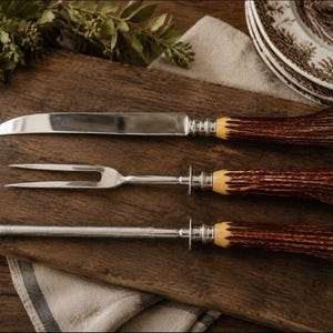 May include: A three-piece carving set consisting of a sharp steel knife, a two-pronged carving fork, and a sharpening steel rod. Each utensil features a textured, dark brown antler-style handle with a polished silver-tone metal bolster.