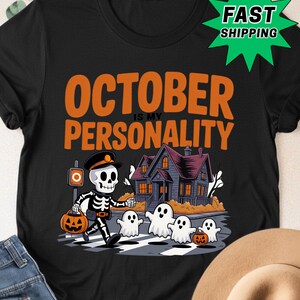 Puede incluir: Una camiseta negra con el texto naranja OCTOBER IS MY PERSONALITY sobre un gráfico de un esqueleto con gorra y un cubo de calabaza, caminando con cuatro fantasmas blancos frente a una casa oscura en una calle con paso de cebra.