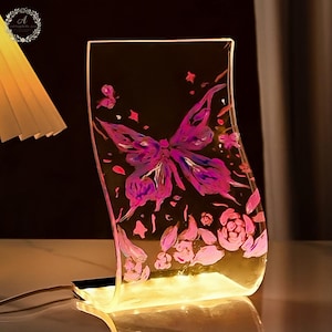 May include: A curved, transparent acrylic lamp featuring a vibrant purple and pink butterfly illustration surrounded by small floral accents. The base emits a warm, golden light that illuminates the butterfly design from below, creating a glowing effect.