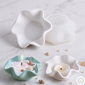 May include: A collection of star-shaped silicone molds and finished candle holders. Two white molds are shown alongside a light green candle holder filled with white sand and seashells, and a white candle holder containing a lit tea light candle.