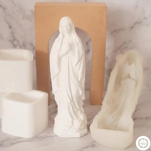 May include: A white silicone mold of the Virgin Mary standing with hands in prayer, accompanied by a separate white square silicone mold and a tan arch-shaped support piece. The mold features detailed drapery and a base for casting figures.