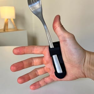Op de afbeelding: Een roestvrijstalen vork bevestigd aan de handpalm met een zwarte, flexibele, U-vormige siliconen band. Het bestek wordt op zijn plaats gehouden door de band die om de handpalm loopt, waardoor de vork omhoog uit de hand steekt voor grip.
