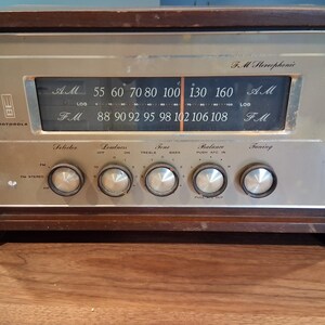 May include: A vintage Motorola FM Stereophonic radio with a dark brown wooden cabinet and a brushed metal front panel. The display features AM and FM frequency scales with a needle indicator. Five circular silver knobs control selector, loudness, tone, balance, and tuning functions.