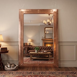 May include: A large rectangular floor mirror featuring a wide, hammered copper frame with a metallic finish. The mirror reflects a living room interior with a brown leather sofa, a wooden coffee table, a fireplace, and a chandelier.