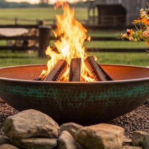 Puede incluir: Una hoguera grande y redonda en forma de cuenco con un interior de cobre martillado y un exterior con pátina verde azulada desgastada. La hoguera contiene troncos ardiendo con llamas de color naranja brillante, rodeada de piedras grises.