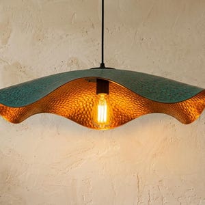May include: A pendant light fixture featuring a wide, wavy-edged shade with a textured teal exterior and a hammered gold interior finish. The lamp is suspended by a black cord and contains a single glowing Edison-style light bulb in the center.
