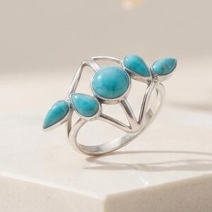 May include: A silver ring featuring a central round turquoise stone flanked by four smaller, teardrop-shaped turquoise stones arranged in a symmetrical, open-work metal design. The band is polished silver, showcasing a clean, structural aesthetic.