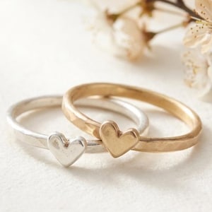 Può includere: Due sottili anelli impilabili, uno in argento e uno in oro, ciascuno caratterizzato da un piccolo ciondolo a forma di cuore centrale. Le fasce presentano una finitura leggermente martellata e testurizzata, con cuori lisci e pieni.