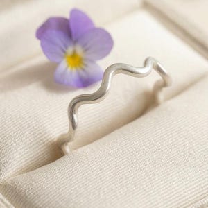 Puede incluir: Un anillo de plata con un diseño de banda ondulada continua. El anillo está colocado en posición vertical junto a una pequeña flor de pensamiento de color violeta claro con un centro amarillo. El metal tiene un acabado liso y mate.