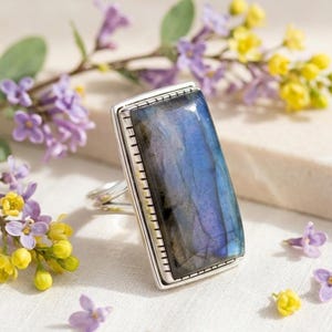 Puede incluir: Un anillo con una piedra preciosa de labradorita rectangular engastada en un marco de metal plateado con un borde dentado. La piedra muestra tonos azules y grises iridiscentes con una superficie lisa y pulida, colocada verticalmente sobre una superficie clara.