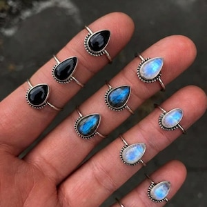 Puede incluir: Una colección de ocho anillos de plata con piedras preciosas en forma de lágrima. Los anillos incluyen piedras de ónice negro, labradorita azul iridiscente y piedras de luna translúcidas, engastadas en bandas de plata con textura metálica.