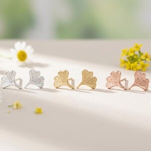 May include: Three pairs of stud earrings shaped like ginkgo leaves, arranged in a row. The first pair is silver, the second pair is gold, and the third pair is rose gold. Each earring features a textured surface with fine, radiating lines.